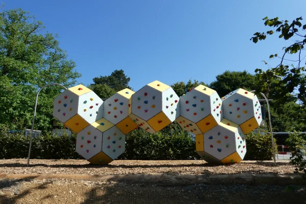 vue-frontale-paysage-grimpe-cube-bloc Installation modulaire de bloc et de grimpe en extérieur avec corps de grimpe polygonaux, prises d’escalade colorées et protection contre les chutes en copeaux de bois.