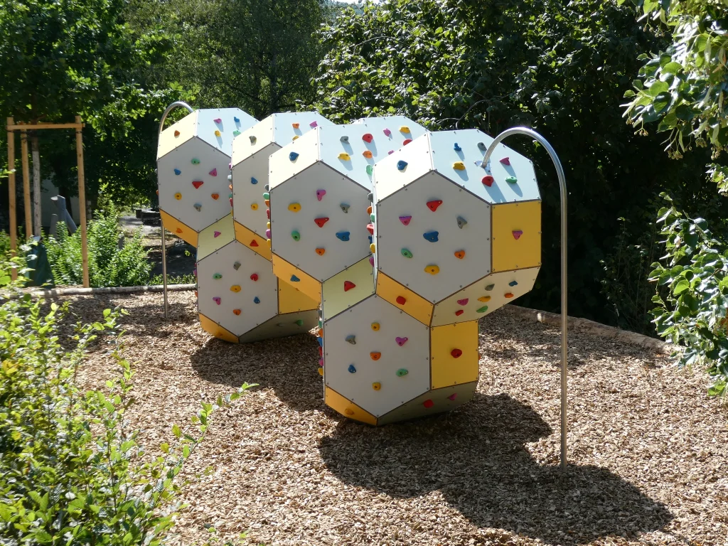 Installation modulaire de bloc et de grimpe en extérieur avec corps de grimpe polygonaux, prises d’escalade colorées et protection contre les chutes en copeaux de bois.